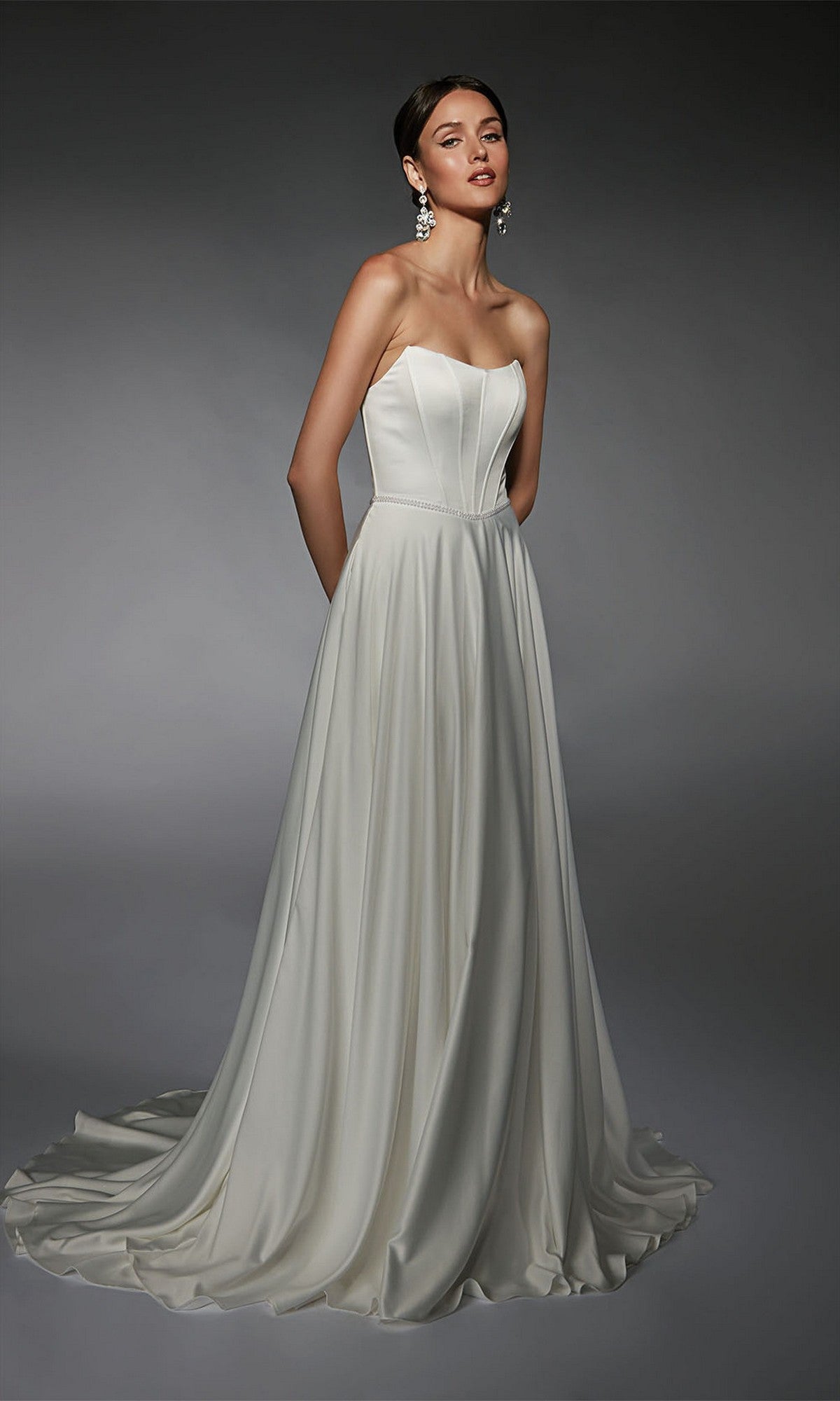 A woman poses against a gray background in the Alyce 7110 Long White Dress, featuring a strapless fitted bodice and flowing skirt, styled with statement earrings for an elegant look.
